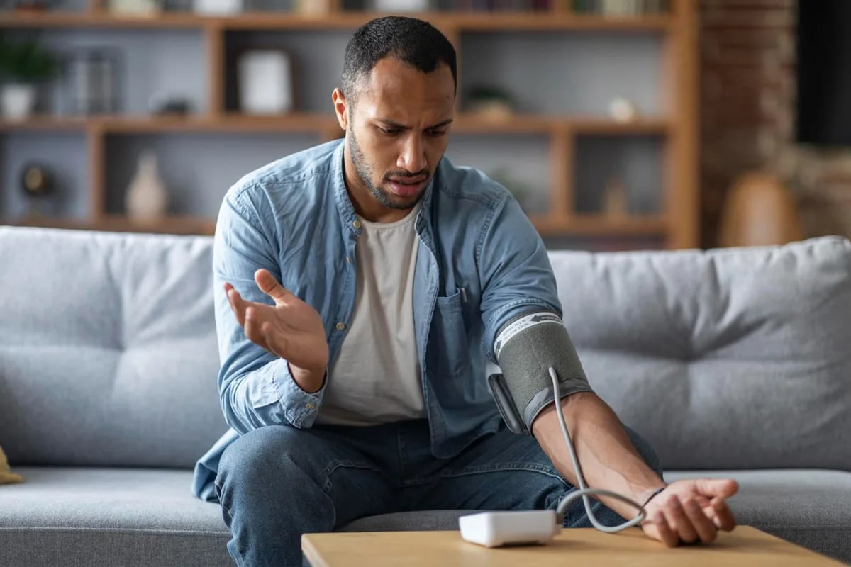 Adult measuring blood pressure at home with upper arm cuff, seated at kitchen table, feet flat, arm supported properly.